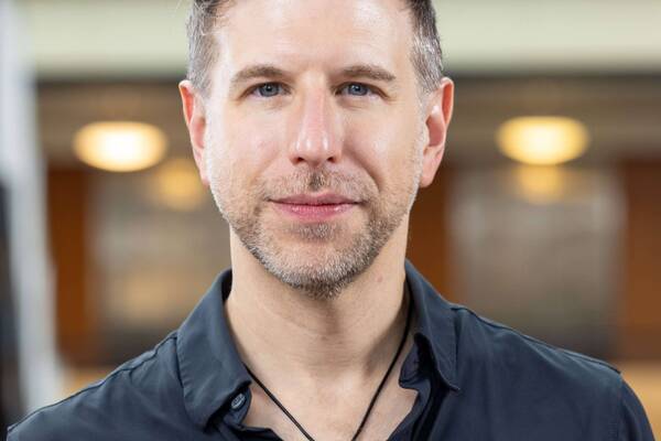 A man with short brown hair and stubble wears a dark shirt and a snake pendant.