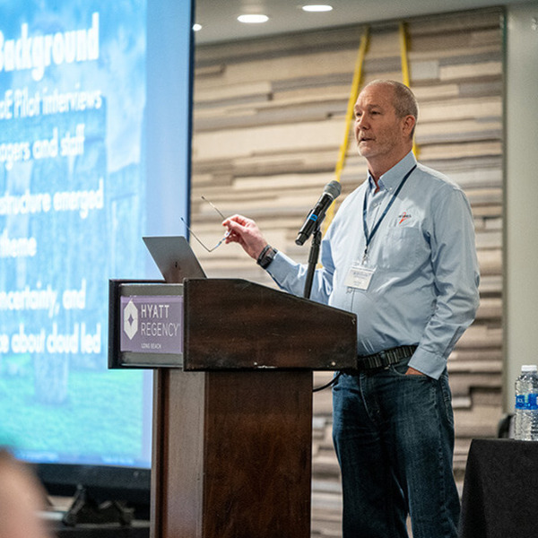 A man in a light blue shirt and jeans speaks at a Hyatt Regency podium, holding glasses. A screen displays "Cloud infrastructure" slides.