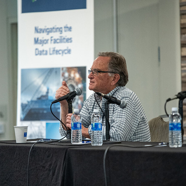 A man sits at a table at the front of a room, with a microphone and water bottles, ready to speak.