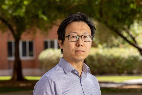 A man with dark hair, black glasses, and a light blue striped shirt looks forward in front of a brick building and trees.